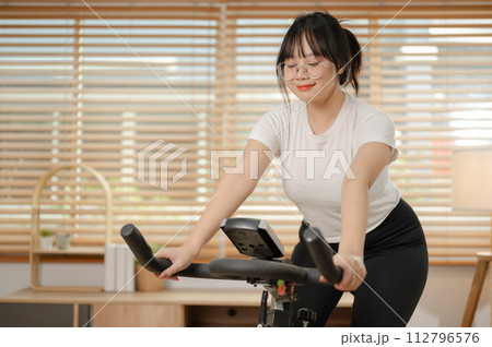 A strong Asian woman in sportswear is exercising on a fitness bike, doing indoor cardio at home. 112796576