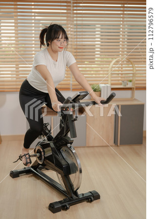 A strong Asian woman in sportswear is exercising on a fitness bike, doing indoor cardio at home. A strong Asian woman in sportswear is exercising on a fitness bike, doing indoor cardio at home. 112796579