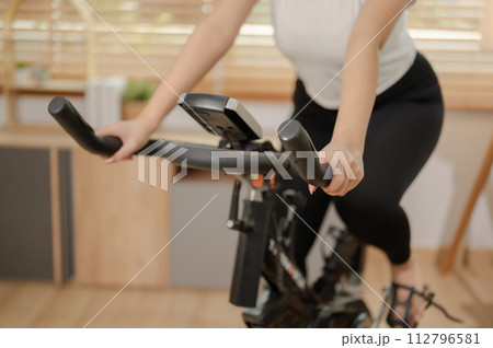 A cropped image of a woman in sportswear exercising on a fitness bike, doing indoor cardio at home. A cropped image of a woman in sportswear exercising on a fitness bike, doing indoor cardio at home. 112796581