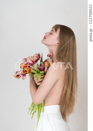 Half naked woman with her eyes closed holds bouquet of spring tulips with her hands and covers chest with flowers. Young Caucasian blonde woman with long hair. Side view 112798022