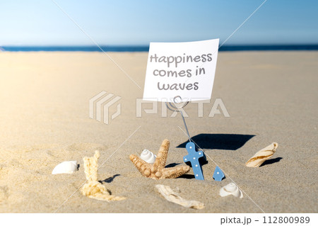 HAPPINESS COMES IN WAVES text on paper greeting card in anchor paper holder and starfish seashell summer vacation decor. Sandy beach sun coast. Holiday concept postcard. Getting away Travel 112800989