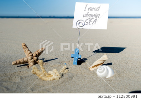 I NEED VITAMIN SEA text on paper greeting card in anchor paper holder and starfish seashell summer vacation decor. Sandy beach sun coast. Holiday concept postcard. Getting away I NEED VITAMIN SEA text on paper greeting card in anchor paper holder and starfish seashell summer vacation decor. Sandy beach sun coast. Holiday concept postcard. Getting away 112800991