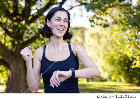 Close-up photo of happy young woman doing sports and jogging in park, looking at smart watch and showing success gesture with hand, rejoicing in victory. Close-up photo of happy young woman doing sports and jogging in park, looking at smart watch and showing success gesture with hand, rejoicing in victory. 112801015