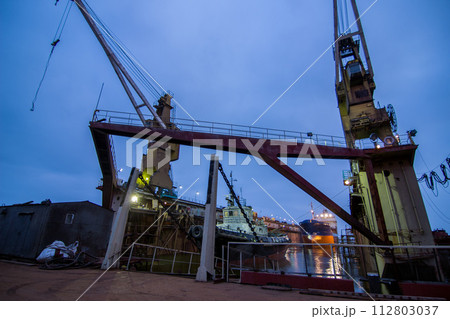 Industrial shipyard at night, floating dock with a ship under repair, lit cranes on duty, marine engineering scene. Ship maintenance, maritime transportation industry, dockyard operations depicted. 112803037