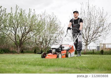 Professional gardener in protective apparel is mowing green grass lawn using modern gasoline cordless lawnmower at backyard. Seasonal landscaping design work. Blooming trees on background 112803608