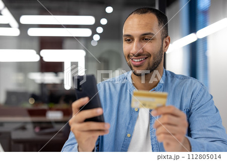 Confident young man holds a credit card and smartphone, shopping online with a smiling expression in a modern office setting. 112805014