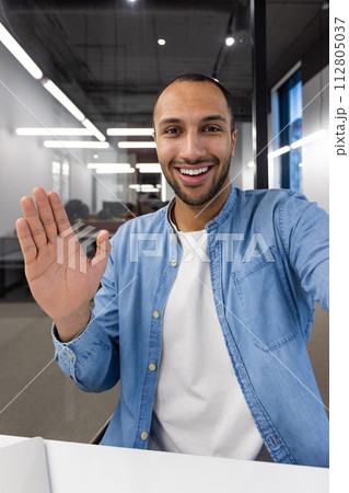 A young, cheerful businessman greets colleagues with a wave during a virtual meeting in a modern office setting. A young, cheerful businessman greets colleagues with a wave during a virtual meeting in a modern office setting. 112805037