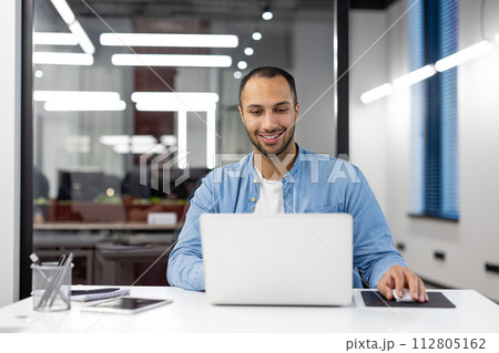 Smiling young professional man using laptop at his desk in a well-lit modern office, showing productivity and comfort. Smiling young professional man using laptop at his desk in a well-lit modern office, showing productivity and comfort. 112805162