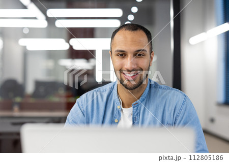 A cheerful young man with a friendly smile working on a laptop in a well-lit contemporary office setting. 112805186