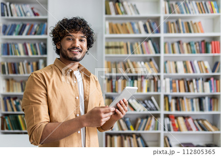 A cheerful young adult male holds a tablet and smiles confidently in a library with bookshelves in the background. 112805365