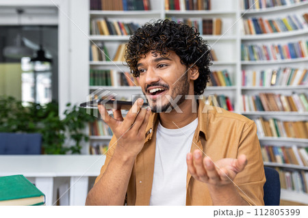 Cheerful young man with curly hair speaks into his smartphone using voice command in a library setting. Cheerful young man with curly hair speaks into his smartphone using voice command in a library setting. 112805399