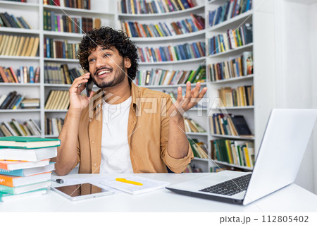 Happy young man with curly hair makes a phone call while working on his laptop in a home office with bookshelves in the background. 112805402