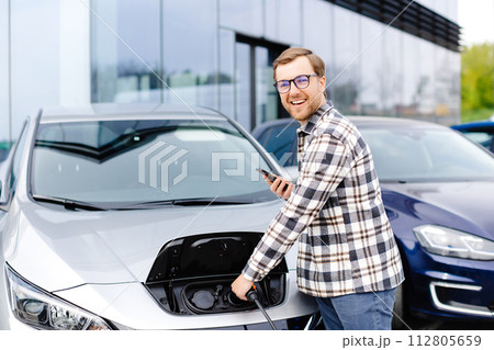 Young man plugging charging cable into the car socket. Electric car charging concept Young man plugging charging cable into the car socket. Electric car charging concept 112805659