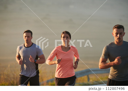 A group of friends, athletes, and joggers embrace the early morning hours as they run through the misty dawn, energized by the rising sun and surrounded by the tranquil beauty of nature A group of friends, athletes, and joggers embrace the early morning hours as they run through the misty dawn, energized by the rising sun and surrounded by the tranquil beauty of nature 112807496