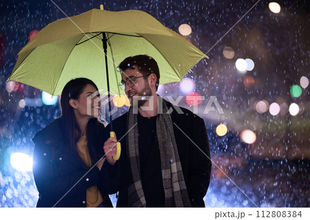 In the romantic ambiance of a rainy night, a happy couple walks through the city, sharing tender moments under a yellow umbrella, surrounded by the glistening glow of urban lights 112808384