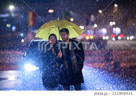In the romantic ambiance of a rainy night, a happy couple walks through the city, sharing tender moments under a yellow umbrella, surrounded by the glistening glow of urban lights 112808435