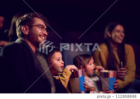 A modern family enjoys quality time together at the cinema, indulging in popcorn while watching a movie with their children 112808698