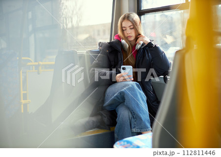 Young girl with headphones sitting in modern tram and looking on mobile phone, listening to music in headphones. Blurred city streets on background Young girl with headphones sitting in modern tram and looking on mobile phone, listening to music in headphones. Blurred city streets on background 112811446