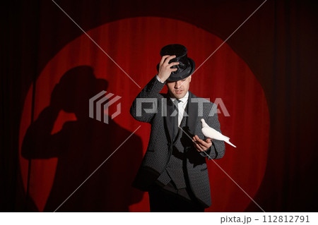 Portrait of magician with pigeon standing over red drapery in spotlight Portrait of magician with pigeon standing over red drapery in spotlight 112812791