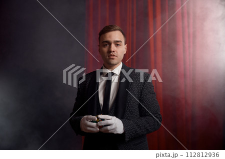 Magician wearing suit standing with cane on stage decorated red velvet curtain Magician wearing suit standing with cane on stage decorated red velvet curtain 112812936