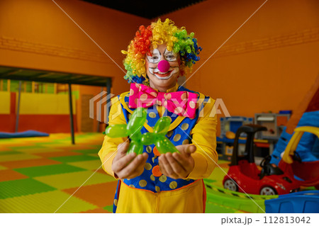 Funny clown holding balloon dog standing over playroom background Funny clown holding balloon dog standing over playroom background 112813042