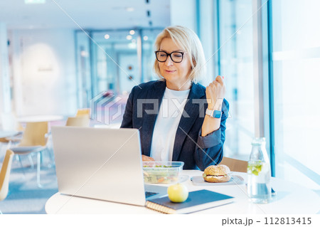 Middle-aged business woman working on laptop and having healthy lunch at working place, office cafe. Alternative office lifestyle female working in video call conference during lunch time. Modern job Middle-aged business woman working on laptop and having healthy lunch at working place, office cafe. Alternative office lifestyle female working in video call conference during lunch time. Modern job 112813415