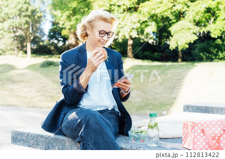 Middle-aged business woman eating sandwich burger with salad and using phone sitting on bench in the park while having lunch break. Remote work,business, freelance,blogging,social media concept. 112813422