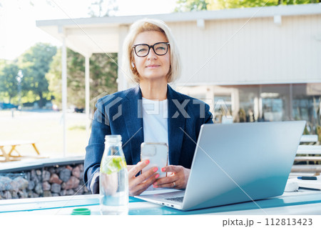 Smiling 50's stylish, confident mature business woman, middle aged lady using wireless ear buds, phone and laptop, working remotely in park. Caucasian woman make a video call, study, work online 112813423