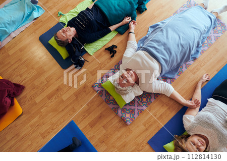 Top view of group of senior women engage in various yoga exercises, including neck, back, and leg stretches, under the guidance of a trainer in a sunlit space, promoting well-being and harmony 112814130
