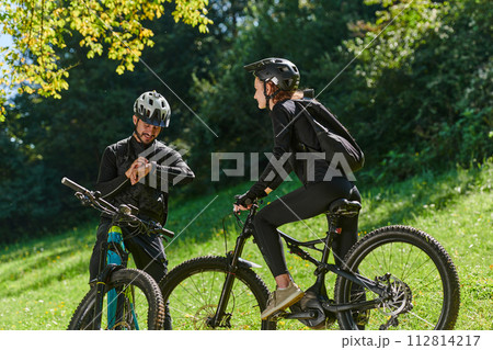 A sweet couple, equipped with bicycles and engrossed in coordinating their journey, checks their GPS mobile and watches while planning scenic routes in the park, seamlessly blending technology and 112814217