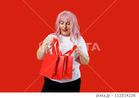Portrait of an excited beautiful woman holding red shopping bags isolated over red background. Black Friday, sale, buy day concept. Pink hair, studio shoot 112814226