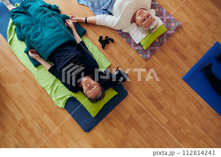Top view of group of senior women engage in various yoga exercises, including neck, back, and leg stretches, under the guidance of a trainer in a sunlit space, promoting well-being and harmony 112814241