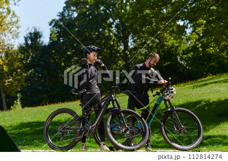 A sweet couple, equipped with bicycles and engrossed in coordinating their journey, checks their GPS mobile and watches while planning scenic routes in the park, seamlessly blending technology and 112814274