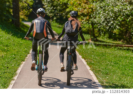 A blissful couple, adorned in professional cycling gear, enjoys a romantic bicycle ride through a park, surrounded by modern natural attractions, radiating love and happiness 112814303