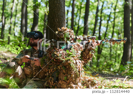 A skilled sniper and a soldier operating a drone with VR goggles strategize and observe the military action while concealed in the forest 112814440
