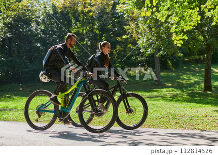 A blissful couple, adorned in professional cycling gear, enjoys a romantic bicycle ride through a park, surrounded by modern natural attractions, radiating love and happiness 112814526