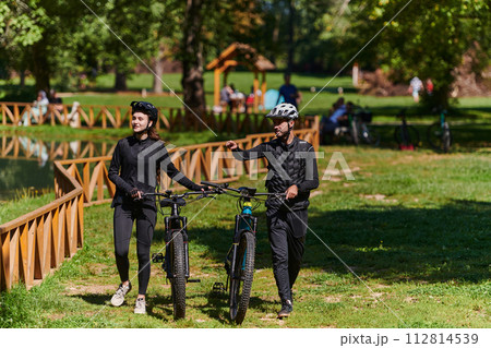 A blissful couple, adorned in professional cycling gear, enjoys a romantic bicycle ride through a park, surrounded by modern natural attractions, radiating love and happiness 112814539