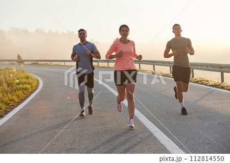 A group of friends, athletes, and joggers embrace the early morning hours as they run through the misty dawn, energized by the rising sun and surrounded by the tranquil beauty of nature 112814550