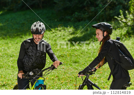 A blissful couple, adorned in professional cycling gear, enjoys a romantic bicycle ride through a park, surrounded by modern natural attractions, radiating love and happiness 112814819