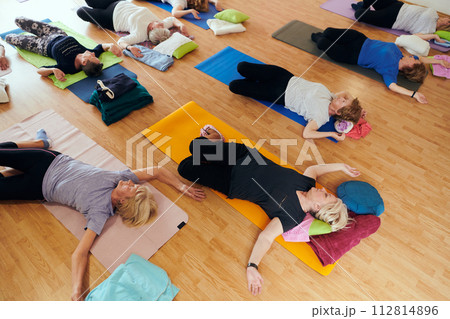 A group of senior women engage in various yoga exercises, including neck, back, and leg stretches, under the guidance of a trainer in a sunlit space, promoting well-being and harmony 112814896