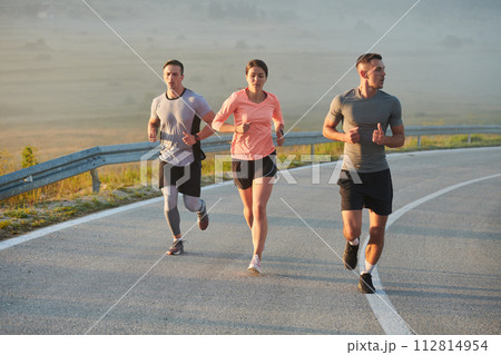 A group of friends, athletes, and joggers embrace the early morning hours as they run through the misty dawn, energized by the rising sun and surrounded by the tranquil beauty of nature A group of friends, athletes, and joggers embrace the early morning hours as they run through the misty dawn, energized by the rising sun and surrounded by the tranquil beauty of nature 112814954