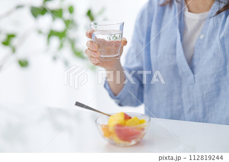 水のグラスを手にフルーツを食べるミドル女性の手元 112819244