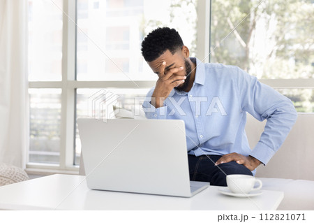 Exhausted frustrated African remote employee man sitting at laptop 112821071