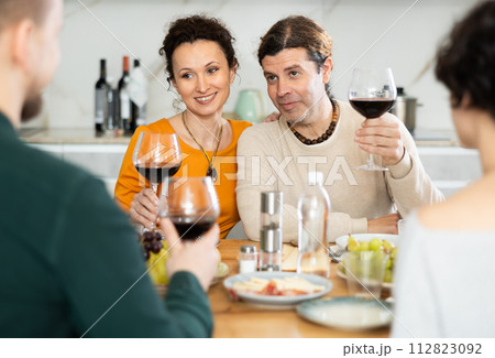 Group of joyful individuals are celebrating a holiday, enjoying drinks and conversation around table in their home 112823092