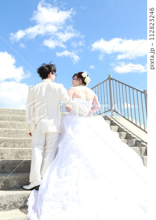 青空をバックにロケ撮影、ウエディングドレス姿の新婦様と白タキシードの新郎様 112823246