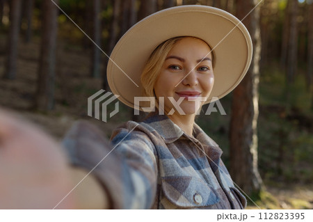 Attractive happy young hipster woman wearing a stylish hat in a fashionable one stands in the park and takes a selfie on the phone. Taking selfies in nature. Close-up Attractive happy young hipster woman wearing a stylish hat in a fashionable one stands in the park and takes a selfie on the phone. Taking selfies in nature. Close-up 112823395