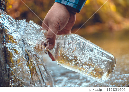A hand brings a transparent bottle to a spring of drinking water. A man collects mineral water from a spring in the autumn forest. The traveler stopped at a spring to drink and collect clean water. 112831403