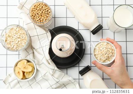 Bottles and glass with alternative milk, towel, hand and bowls with ingredients on white background, top view 112835631