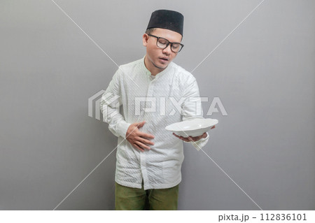 Asian Muslim man wearing a koko shirt and peci with shades of the fasting month, holding back hunger and plate while waiting for the time to break the fast, isolated on a gray background 112836101
