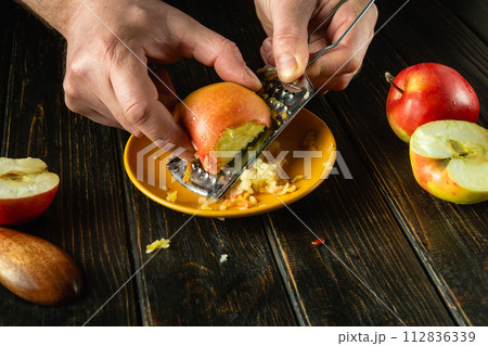 Preparing a dietary dish with apples. Chef grates a raw apple with a grater on the kitchen table. Advertising space on a black background Preparing a dietary dish with apples. Chef grates a raw apple with a grater on the kitchen table. Advertising space on a black background 112836339